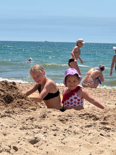 The image depicts a group of people enjoying a sunny day at the beach in Vityazevo, Russia. The scene is captured from a low angle, emphasizing the sand in the foreground and the shimmering ocean in the background.

In the foreground, a young girl with blonde hair, wearing a purple hat and a strawberry-print swimsuit, is smiling at the camera. She is partially covered by sand. To her left, another young girl with blonde hair and a black bikini top is looking towards the water.

Further back, a woman in a leopard-print bikini and black bottoms is seen from the waist down, reaching towards the water. Behind her, a woman in a white and yellow bikini is squatting down, holding a blue towel.

The water is calm with gentle waves lapping the sandy shore. The sky is a clear, bright blue. The overall atmosphere is one of leisure and enjoyment on a summer day. No visible text is present in the image.
