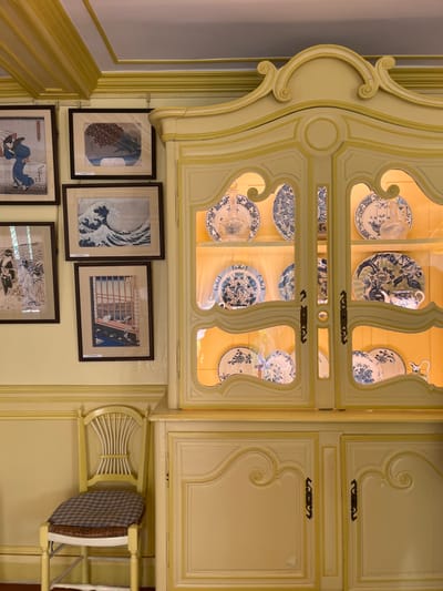 The image shows a yellow, ornate, two-door cabinet filled with blue and white porcelain plates, in a room with pale yellow walls. The room also contains 5 framed pictures, all of which are Japanese woodblock prints, and one wooden chair with a checkered seat cushion. The cabinet has a long, slightly curved top with ornate floral patterns. The lower half of the cabinet has two doors with two simple, horizontal handles. The cabinet is located in the dining room of the house of Claude Monet in the town of Giverny, France, located at coordinates 49.075, 1.534.  The house is known for its gardens, which were the subject of many of Monet's paintings.