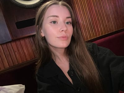A female subject is depicted in a close-up, chest-up selfie, centrally positioned within an indoor setting. She has long, straight, light brown hair, fair skin, and light-colored eyes, looking directly at the camera with a neutral expression and slightly parted lips. She wears small gold hoop earrings and a dark-colored, likely black, collared shirt or jacket. A thin silver necklace with a small clear pendant is visible on her chest. The background consists of dark wood paneling with horizontal grooves on the left, partially obscuring a large, dark circular object (possibly a speaker) in the upper left. To the right, another section of reddish-brown wood paneling features vertical grooves, beneath which a dark red upholstered bench or booth seat is visible. A white object, possibly paper or fabric, is partially present in the bottom left corner. The lighting is dim and warm, characteristic of an evening establishment. The specific location within 마포구, South Korea, is not discernible from the visual evidence presented in the image.