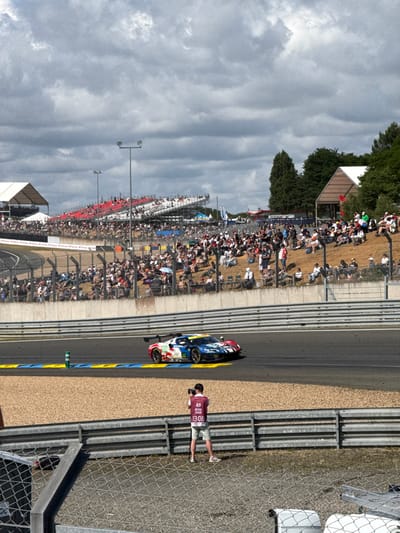 A race car speeds along a track lined with spectators in bleachers. A photographer wearing a red vest stands behind a chain-link fence, capturing the action. The race track has blue and yellow curbing. Light poles stand along the sidelines of the race course. The sky is cloudy.