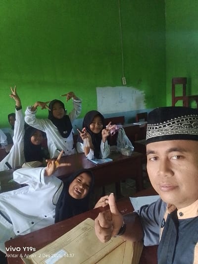 A man wearing a Muslim prayer cap takes a selfie with a group of students in a classroom. The students, all girls wearing hijabs and white uniforms, are making hand gestures, smiling, and posing for the camera. The classroom has green walls and wooden desks, with a whiteboard visible in the background. One student is lying on the desk and giving a thumbs up. A plastic bag of food or drink sits on one of the desks. The image includes a watermark at the bottom left.