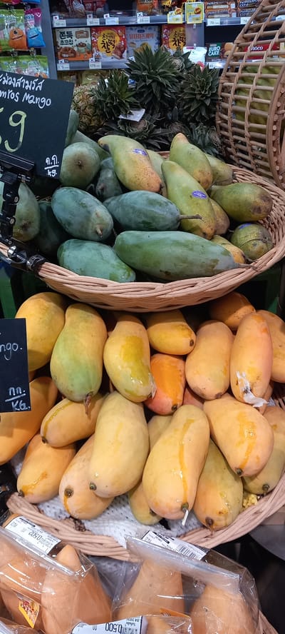 This image captures a vibrant section of a grocery store or market, likely in Moo 1, Thailand, featuring an abundant display of fresh mangoes and packaged goods.

In the foreground, two large, woven baskets are filled with mangoes. The basket on the left contains a mix of green and partially ripened yellow-green mangoes, with several spiky pineapples nestled behind them. A black pricing sign in front of this basket reads "ม่วงสามรัส" (Mang Sam Rus) and "mros Mango," indicating the variety, along with "ก.ก. / kg." The number '9' is partially visible on the sign. The basket on the right is overflowing with bright yellow and orange ripe mangoes, some exhibiting sticky residue or natural juices on their skin. Several of these ripe mangoes are individually packaged in clear plastic, with visible labels showing "น้ําหนักสุทธิ 0.502 กก." (Net weight 0.502 kg) and "วันที่ผลิต 6.04.2020" (Production date 06.04.2020) on one, and "0.500 กก" (0.500 kg) on another.

To the left of the fruit display, shelves are stocked with various packaged snacks and biscuits. Visible product names include "チョコチップ" (Choco Chip), "チョコ&コーヒービスケット" (Choco & Coffee Biscuit), "초코파이" (Choco Pie), "유자치즈케익" (Yuja Cheese Cake), and "EURO CAKE." Prices in Thai Baht are displayed on shelf labels, such as "70," "55," "50," "175," and "45," with some "Sale" signs. The scene portrays a lively retail environment, offering both fresh local produce and a variety of international convenience items.