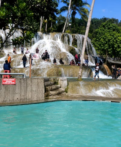 The image shows a tiered waterfall with people interacting with the water. In the foreground, a bright turquoise swimming pool is visible, with a concrete edge and a wooden barrier. A red sign is attached to the wooden barrier, with text in Indonesian that reads "DILARANG BERENANG DAN BERMAIN DI AREA AIR TERJUN" which translates to "SWIMMING AND PLAYING ARE PROHIBITED IN THE WATERFALL AREA".
Behind the pool, the waterfall cascades down a rocky, multi-layered formation. Several individuals are seated on the rocks, some near the water's edge, while others are standing or walking on the wet surfaces. A prominent palm tree trunk is positioned in front of the waterfall. Trees and foliage surround the top of the waterfall, with a clear blue sky visible above. The scene appears to be a public recreational area, likely within the city of Silaiyajae, Indonesia, based on the provided context, featuring a natural waterfall integrated with man-made swimming facilities.