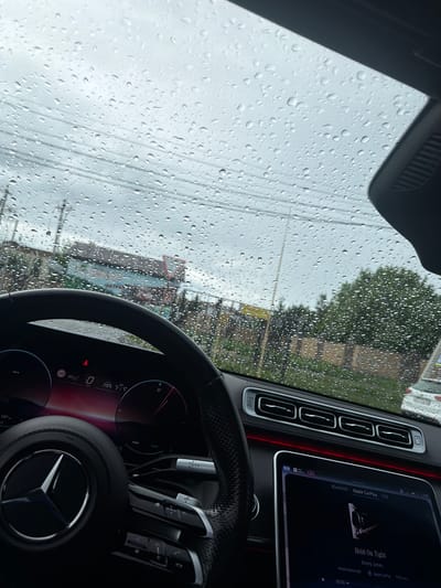 Here's a detailed description of the image:
**Scene:** The image is taken from inside a Mercedes-Benz car, likely in the driver's seat. The windshield is covered in raindrops, obscuring the view of the outside.
**Interior:** The car's interior is visible, including the steering wheel with the Mercedes-Benz logo, the dashboard with digital displays, and the infotainment screen. The infotainment screen shows that the car is connected via Apple CarPlay and playing "Hold On Tight" by Boney James. The car is stationary, indicated by the speedometer reading 0 km/h.
**Exterior:** Through the rain-covered windshield, a street is visible. There are buildings and trees, but the details are obscured by the rain. There are power lines strung across the sky.
**Inferred location:** Though not explicitly stated within the image, it is provided in the prompt that the location is Krasnyi Posyolok, Russia.