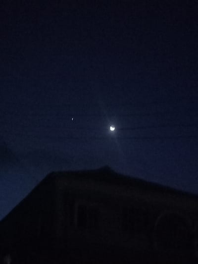 The image shows a crescent moon and a star in a dark sky, with the silhouette of a building in the foreground. The location is Ita Oniyan, Nigeria, at coordinates (latitude: 7.292, longitude: 5.167). No people or objects are visible except for the moon, star, and building. The building is mostly obscured by the darkness of the night. The image appears to have been taken at night. The moon is the most prominent feature in the image, with the star being less visible. The image is taken from a low angle, with the building filling the majority of the frame. The image is dark, with only the moon and star providing light.