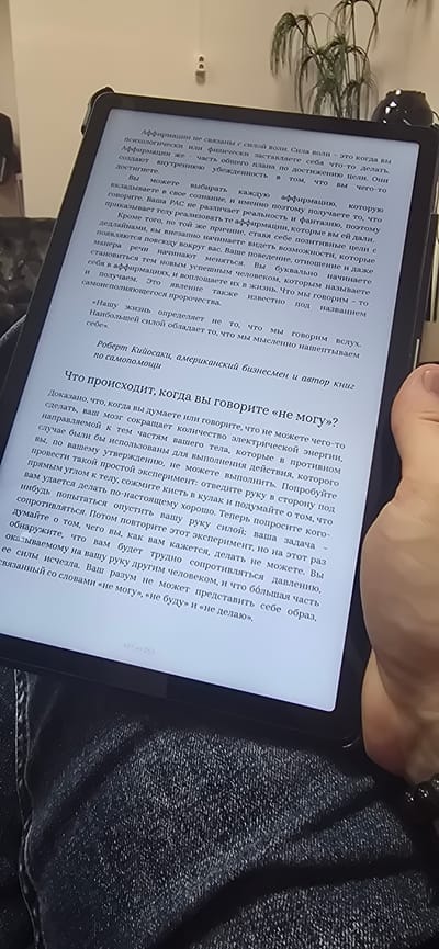 The image shows a close-up of an e-reader device displaying text. The device is held by a person whose hand is partially visible in the lower right corner, wearing a bracelet. The text on the e-reader appears to be from a book discussing affirmations and personal development, with headings like "What happens when you say 'I can't'?" and quotes from Robert Kiyosaki. The background is blurred and shows hints of indoor lighting and possibly furniture. There are no discernible landmarks or elements that would indicate a specific location in Bryansk, Russia. The scene is an indoor, personal setting focused on the act of reading.