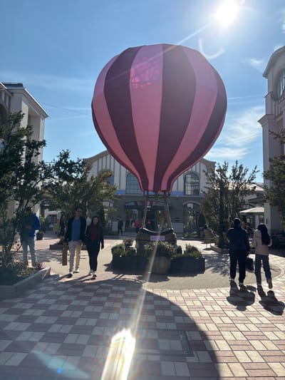 The image portrays an outdoor pedestrian zone within Ingolstadt Village, an outlet shopping complex in Ingolstadt, Germany. The scene is illuminated by bright daylight, with the sun causing a lens flare in the lower left quadrant and casting distinct shadows.
At the center-right, a large, static hot air balloon replica features an envelope with vertical stripes of alternating pink and dark magenta/purple. A wicker basket is attached to its base, which is situated within a circular planter containing green foliage.
The ground is paved with rectangular tiles in an alternating light and dark reddish-brown pattern. Multi-story buildings with light-colored facades, arched windows, and tiled roofs line both sides of the pathway. A "BOSS" storefront sign is visible on one building in the background. Manicured trees and shrubs are strategically planted along the walkways.
Several individuals are present:
* On the far left, a male in a blue top and light-colored pants walks right, partially obscured by vegetation, holding a blue object.
* In the mid-left foreground, a male in a dark jacket and light pants, carrying a brown paper bag, walks alongside a female in a dark top and pants, both moving towards the viewer.
* On the mid-right, two figures, a male in a dark jacket and a female in a light jacket, walk away from the viewer towards the right.
* Additional people are visible in the background, near storefronts and seated at outdoor tables.
The sky is clear blue with minimal wispy clouds.