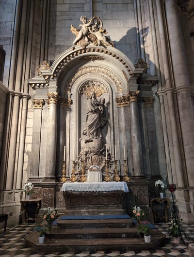 This image shows an interior view of a church altar. The altar is elaborate, with a statue of the Virgin Mary holding the infant Jesus at its center. The statue is set within a large, ornate niche framed by columns and topped with gilded architectural details. Two cherubs are depicted above the niche. The altar itself is draped with a white lace cloth. In front of the altar are several steps, leading down to a floor with a checkered pattern. Various decorative elements, including candle holders and vases of flowers, are placed around the altar and on the steps. The church walls are constructed of stone, with tall, fluted columns visible. The lighting suggests the image was taken indoors, likely in the daytime given the presence of natural light and shadows. The scene is a religious one, common in churches and cathedrals. While the image is identified as being in Urvillers, France, this particular altar cannot be definitively identified as belonging to a specific church in that location without further information.