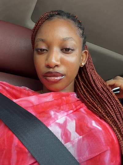 The scene depicts a female individual seated within a vehicle in Ikota, Nigeria. Her upper torso and head are visible in a close-up, angled shot. She has medium brown skin, and her hair is styled in numerous small, reddish-brown box braids that extend over her shoulders. Her eyes are partially closed or downcast. Her lips are coated with a dark red, glossy substance. A small, spherical gold stud earring is visible on her right earlobe. She is wearing a top or dress made of a sheer, fine-mesh fabric, exhibiting a red, pink, and white tie-dye or mottled pattern. A dark grey seatbelt is fastened across her chest, running diagonally from the upper left to the lower right of the frame. The immediate background shows a dark reddish-brown upholstered surface to her left and a lighter, possibly grey or beige, ceiling or upper interior panel to her right. A metallic-colored object is faintly visible on the far right.