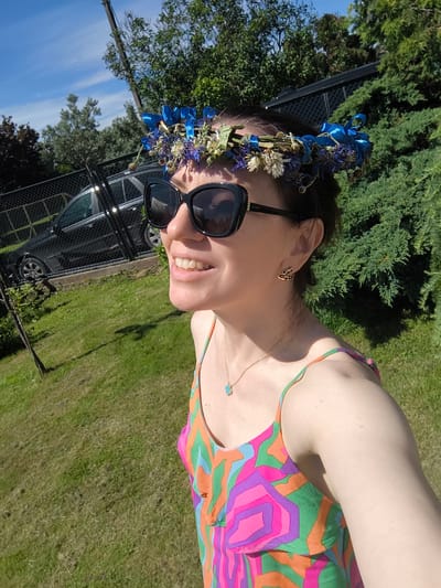 The image shows a woman wearing a floral crown and sunglasses, looking up and to the left. She has a bright smile. She is wearing a colorful, abstract-patterned dress and a delicate necklace with a small turquoise pendant. She also has small, gold-colored earrings. The woman is outdoors in a grassy area with trees and a fence in the background. A car is visible behind the fence. The sun is shining brightly, suggesting it is a clear day. The overall impression is of a cheerful and summery scene, possibly indicating a celebration or outdoor activity.