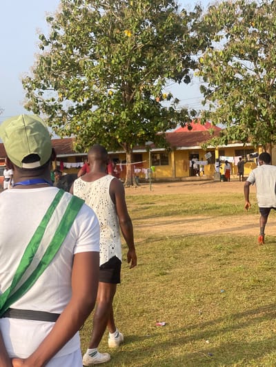 In Emure-Ekiti, Nigeria, the image captures an outdoor scene with several individuals. In the foreground, the back of a person wearing a white t-shirt and a light green cap is visible. A green and white sash is draped across their chest. To their right, another person, viewed from the back, wears a white tank top with a textured pattern and dark shorts. A third person, on the right side of the frame, is seen from the back, wearing a gray t-shirt and dark shorts, with one leg bent as if in motion. In the background, a yellow building with a red roof is visible, with clothes hanging on a line in front of it. Several other individuals are present near the building, some standing and some appearing to be walking. The environment is grassy with dirt paths. The sky is a pale blue.