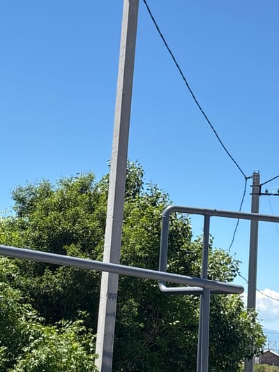 The image depicts a bright, sunny day with a clear blue sky. In the foreground, a grey metal railing with curved sections is visible. Behind the railing, lush green trees dominate the landscape. Two tall, grey concrete utility poles stand vertically, one positioned to the left of center and another to the right, partially obscured by the trees and railing. Wires are suspended between these poles and extend upwards and across the frame. The overall scene suggests an outdoor environment, possibly a park or a residential area with utility infrastructure, located in Jrarat, Armenia.