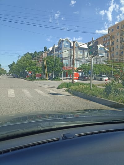 The image shows a street scene in Izberbash, Russia. In the foreground, the hood and dashboard of a vehicle are visible, suggesting the photo was taken from the driver's perspective. The road ahead has a pedestrian crossing marked by white stripes. To the right of the road, a traffic light is visible, currently displaying a green signal. Beyond the road, there are several buildings, including a multi-story apartment building and other structures with a modern architectural style. Trees and green vegetation line the sides of the road and surround the buildings. Signs are visible, one of which appears to advertise a business named "РУСЛАН" (Ruslan) with the text "МЫ ЖДЕМ ВАС" (We are waiting for you). Cars are parked and driving on the street, indicating an active urban environment. The sky is blue with scattered white clouds, suggesting fair weather.