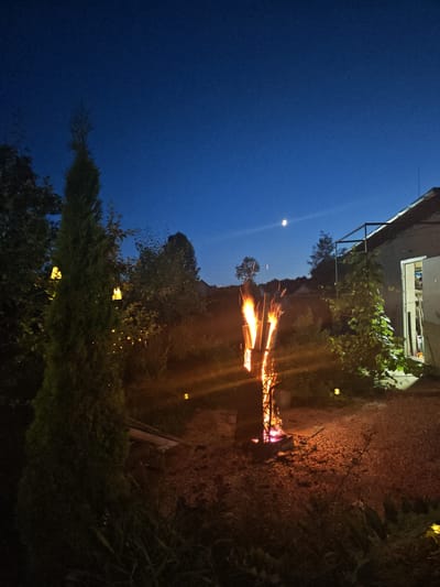 An outdoor scene at dusk or night, likely in a backyard or garden area of Надежда Снт, Russia. The sky is a deep blue, with a sliver of moon visible. In the foreground, a tall, conical evergreen tree stands to the left, decorated with several yellow, lantern-like lights. To the right of the tree, a metal fire pit is actively burning, emitting bright orange flames and sparks. Beyond the fire pit, a simple, light-colored building with an open doorway is visible on the right side of the frame. The ground is covered with gravel and some low-lying vegetation. The overall ambiance is one of a peaceful evening gathering or relaxation.