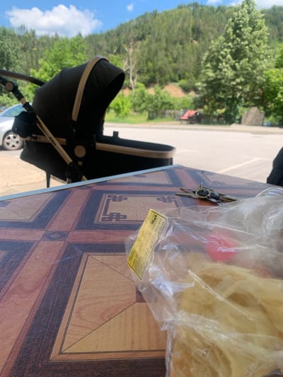 The image shows a close-up view of a table with a wood-grain pattern. On the table, there are a few objects, including a set of keys with a black fob, and a clear plastic bag containing what appears to be food. A yellow label is attached to the plastic bag with some text in Bulgarian. Partially visible in the background is a black baby stroller, suggesting the scene might be outdoors, possibly in a park or a residential area. The background shows lush green trees and a bright blue sky with some clouds, indicating it's daytime with pleasant weather. The overall impression is a casual outdoor setting, perhaps a picnic or a break during a walk. The Bulgarian text on the label mentions "БАНИЦА" (Banitsa), a traditional Bulgarian pastry, and lists ingredients and storage instructions. The word "Albe" is also visible on the stroller. The scene evokes a sense of a relaxed, everyday moment.