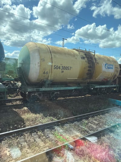 The image shows a train car with a large cylindrical tank, painted yellow and light green at the ends. The car is on a railway track, with gravel and sparse vegetation on either side. In the background, there are other train cars, electrical wires, and a cloudy blue sky. There are markings on the tank car, including the number "504 30057" and a logo that appears to be "GAZPROMTRANS". The train car is likely at a railyard or station in the city of Rossosh, Russia.