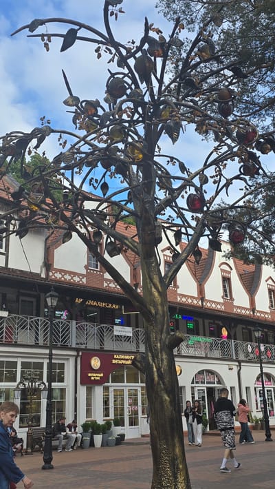 In Svetlogorsk, Russia, an artistic metal tree sculpture with metallic leaves and fruit stands in a paved pedestrian area. Behind the sculpture, a two-story building features arched windows, red-tiled roofs, and multiple shop entrances with signage in Russian. The lower level of the building houses businesses such as "ART SPA Aesthetic Studio," "Kaliningrad Amber Boutique," and other shops. Several people are visible in the scene, including a woman walking towards the left foreground, a group of people sitting on a bench near the entrance of a shop, and individuals walking past the shops in the background. The sky is cloudy.