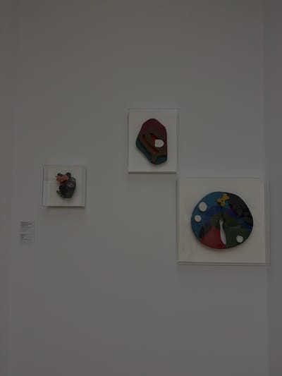 Three framed artworks are displayed on a plain white wall, typical of an art gallery setting. The art pieces are abstract and colorful. On the left, a small, square frame contains a dark gray and pink sculptural piece. Below this artwork, two white placards are mounted on the wall, likely providing information about the art. In the center, a rectangular frame holds another abstract artwork with layered shapes in shades of red, green, blue, and brown, with a white oval shape in the middle. To the right, a larger square frame displays a circular artwork composed of various colored segments, including blue, green, and red, with white circles and a yellow abstract shape near the top. The overall impression is one of a clean, minimalist exhibition space. This scene is located in the Fondation Beyeler museum in Riehen, a municipality of Basel, Switzerland.