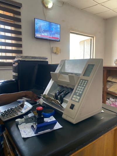 The image depicts an office setting in Jere North, Nigeria, likely a financial institution or administrative office. A person's arm and hand are visible on the left side, interacting with a calculator and possibly feeding paper money into a currency counting machine. The currency counting machine is a prominent object in the foreground, displaying a digital readout and a keypad. Other objects on the desk include a calculator, a stamp with an ink pad, and what appears to be handwritten notes. In the background, a computer tower and monitors are visible, along with blinds on a window and a television screen displaying news content. The environment appears functional, with office furniture and supplies present. The overall scene suggests a typical day's work in such an establishment.