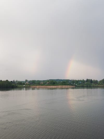 The image depicts a tranquil water scene, likely a lake or wide river, in the vicinity of Анхимово, Russia. The water has gentle ripples across its surface. The shoreline is lined with a mix of trees and reeds, suggesting a natural or semi-natural environment. A few houses are visible among the trees, indicating the presence of a small settlement or village. A distinct rainbow is present in the sky, its arc partially visible. A reflection of the rainbow appears mirrored in the water. The sky is overcast, with a muted, gray tone.