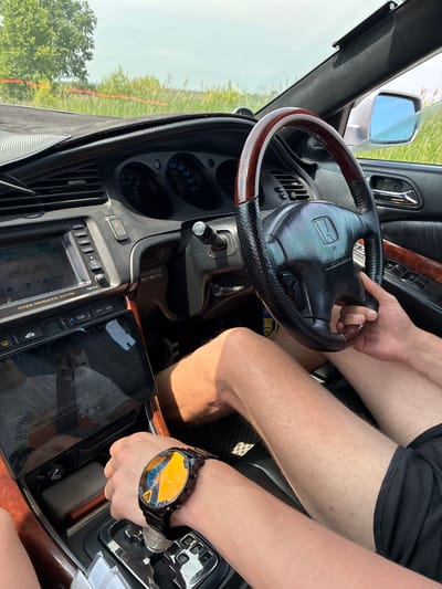 The image shows the interior of a car, likely a Honda, with a person's hands visible. The steering wheel is black with a wood-grain trim. A dashboard with a navigation screen and instrument cluster is also visible. A watch with a yellow and blue face is worn on one of the hands. The car appears to be in motion, as suggested by the slightly blurred background of green vegetation and a bright sky. The setting appears to be rural, with no buildings or urban features visible. The image was taken on a sunny day.