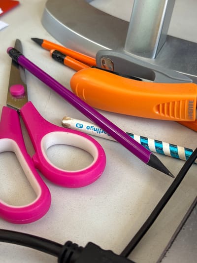 The scene, located in Tayshet, Russia, depicts a static, close-up view of various stationery items and office supplies arranged on a light-colored desk surface. In the foreground, a pair of pink-handled scissors with "STAINLESS STEEL" imprinted on a blade is visible, accompanied by a pink circular stopper. A sharpened purple pencil with a black tip lies across the scissors and other items. Two sharpened orange pencils are situated in the upper-middle section, near a grey stapler with a metallic silver arm. An orange utility knife with a visible "VENTE" logo is positioned to the right of the pencils. Further right, a short white pencil marked "Berlingo HB" and a blue and white striped pencil are present. A large, silver-grey object, likely the base of a monitor, is in the background. A black cable with a dark connector extends into the lower foreground from the right. A small corner of a red object and a piece of white paper are partially visible in the upper left background. No individuals or actions are depicted.