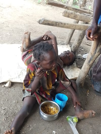 In Otulu, Nigeria, two young children are depicted in an outdoor setting with a dirt ground and wooden structures. One child, sitting in the foreground, is wearing a colorful, patterned shirt and is in the process of eating from a metal bowl with a spoon. A blue cup is placed beside the bowl. This child's legs are visible, and they appear to be sitting on a white sheet or tarp.
Behind the foreground child, another child is lying down on the white sheet. This child is looking towards the viewer. The background features rough wooden beams and posts, suggesting a rustic or unfinished construction. To the right of the children, a person's legs and feet are visible, wearing dark sandals. One foot rests near two green fruits enclosed in a plastic bag.
The scene appears to be in daylight, with natural lighting. The overall impression is one of a humble, possibly impoverished environment. There is no visible text in the image. The children's expressions are not clearly discernible for emotional interpretation.