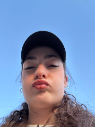 A young woman with curly brown hair wears a black baseball cap against a bright blue sky. She is looking down at the camera, her eyes closed in a relaxed or playful expression. Her lips are slightly pursed. She has delicate silver hoop earrings and appears to be outdoors, possibly tilting her head back to capture the shot.