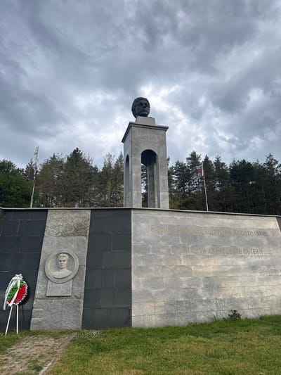 The image captures a monument in Bunovo, Bulgaria, dedicated to Vasil Levski and Panayot Volov, prominent figures in Bulgarian history. The monument consists of a tall, light gray stone tower topped with a bust of Vasil Levski, accompanied by a larger dark gray stone wall. Etched in Cyrillic letters on the stone wall are quotes attributed to Vasil Levski and a reference to Zahariy Stoyanov. A second bust, carved on a light-gray stone disc, depicts Panayot Volov. A wreath with red, green, and white flowers lies at the base of the monument. The Bulgarian flag flies behind the monument. The surroundings include trees and grass. The sky is overcast with heavy gray clouds.