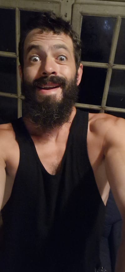 A man with a full beard and dark hair is looking directly at the camera with wide eyes and an open mouth, appearing surprised or excited. He is wearing a black tank top. The background consists of a dark, multi-paned window. The image is a close-up shot of the man's face and upper torso. There is no discernible location in the city of Wattenwil, Switzerland.