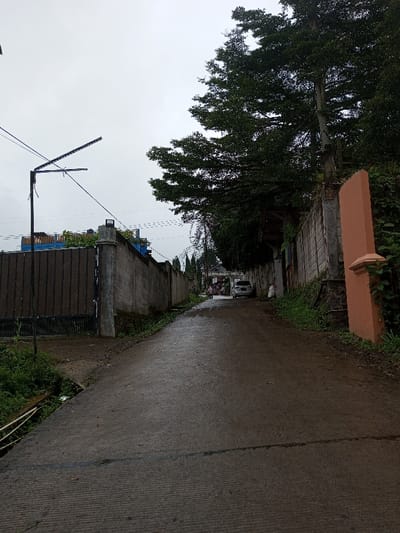 The image displays an uphill, dark gray paved road in YonPhoogla, Bhutan, under an overcast, light gray sky. The road surface appears wet. On the left side of the foreground, a shallow ditch contains green vegetation and light-colored debris. A tall, dark gray metal gate leads to a concrete block wall extending upwards. A black utility pole with a horizontal arm and attached wires stands beside the road. Beyond the wall on the left, a building with a visible blue roof is partially obscured. On the right side of the foreground, green vegetation borders a light orange concrete pillar. This pillar is adjacent to another concrete block wall, which runs parallel to the road. A large, dense green tree with extensive foliage overhangs the right wall, extending into the upper portion of the frame. Further up the road, a white multi-purpose vehicle is parked on the right side. Two individuals, an adult and a child, are visible near the parked vehicle. A white bag or sack is on the ground beside the vehicle. A white gate or archway structure is discernible at the crest of the incline.
