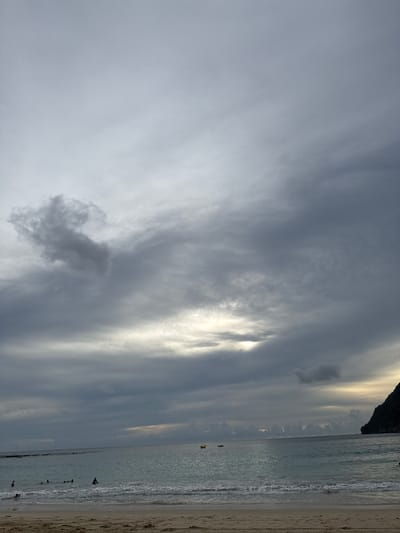 This is a photograph of a beach scene at Meunasahbale, Indonesia, under a cloudy sky. The foreground features a sandy beach with visible footprints. The middle ground shows the ocean with gentle waves breaking near the shore. Several people are in the water, swimming and wading. A yellow buoys is visible in the water. In the distance, a dark, rocky landmass juts out into the sea on the right side of the image. The sky is filled with heavy, grey clouds, with some lighter patches suggesting the presence of the sun behind them.