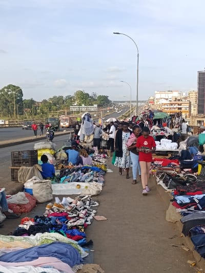 The image depicts an informal outdoor market operating alongside a multi-lane elevated highway in Kiuu ward, Kenya. The foreground features a paved pedestrian walkway densely arranged with merchandise, primarily second-hand clothing and footwear, displayed on the ground, on sheets, and in various piles. Numerous pairs of sneakers and other shoes, alongside folded garments, are visible. Vendors utilize plastic crates and large fabric sacks for display and storage. A green canopy structure is present among the stalls on the right side.
Approximately twenty-five individuals are observed within the frame. A woman wearing a red shirt and light-colored leggings walks towards the viewer, holding an unspecified object, followed by another woman in a patterned dress. Other individuals are dispersed throughout the market area, some kneeling or standing near the merchandise, indicating activities consistent with browsing or vendor operations.
The background shows a multi-lane road with vehicular traffic, including cars, a truck, and a motorcycle. Trees and dense green foliage line the left side of the highway. Multi-story urban buildings are visible on the right side of the highway in the distance. Multiple streetlights with curved poles are positioned along the thoroughfare. The sky above is partly cloudy.