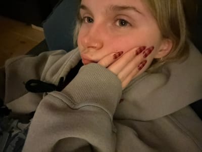 A young woman with blonde hair poses for a selfie, holding a hand up to her face, showcasing long, maroon-colored nails with decorative designs. She wears a light grey hooded sweatshirt with black drawstrings. A silver nose ring is visible. The background is slightly blurred, showing what appears to be a wooden floor and a blue fabric surface.