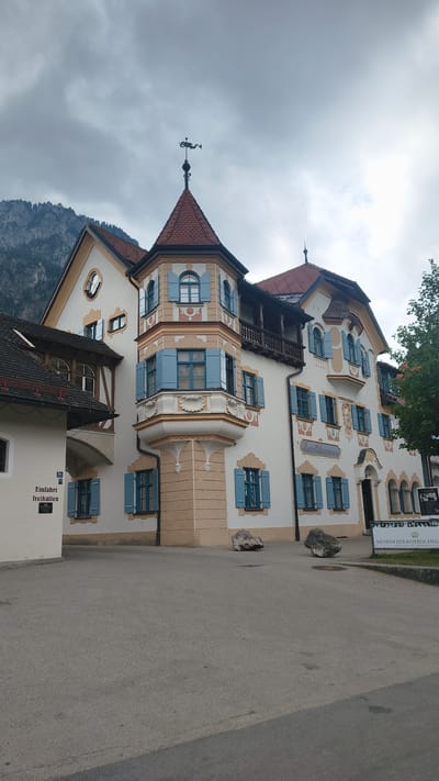 The image shows a three-story building with a unique architectural style in Schwangau, Germany. The building has a white facade with light brown and cream-colored decorative elements. It features numerous windows with bright blue shutters, a prominent turret with a red tiled roof, and a weather vane on top. The building also has a balcony on the second floor. To the left of the main building, there is an attached structure with a dark tiled roof and a sign that reads "Einfahrt freihalten" (Keep entrance clear). In front of the building, there is a paved area that appears to be a parking lot or courtyard, with two large rocks placed on the ground. In the background, mountains can be seen under a cloudy sky. A sign for "MUSEUM DER BAYERISCHEN" is visible on the right side of the image, suggesting the building houses a museum. There are no people visible in the image. The overall scene depicts a historical building in a picturesque, possibly Bavarian, setting.