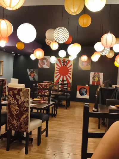 The image depicts the interior of a restaurant, potentially located in Quito, Ecuador, decorated with Japanese-themed art and lanterns. The restaurant has multiple tables and chairs, some of which are occupied by patrons. A large rising sun flag and other pop art-style images of Japanese figures adorn the black walls. The floor is light-colored wood. Lanterns of various sizes and colors hang from the ceiling, providing ambient lighting. There are people seated at some tables, although specific activities are not clearly visible. The scene conveys a sense of dining in a Japanese-inspired setting.
