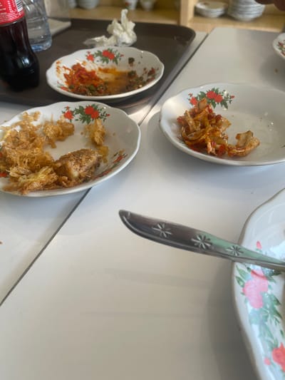 The image depicts a table with leftover food in several white, floral-patterned plates. In the foreground, a fork rests on the table, its handle showing a starburst pattern. To the left, a partially eaten meal is visible in a plate, consisting of crispy fried items and a piece of meat. Another plate to the right contains remnants of what appears to be shredded meat in a sauce. A third plate in the background shows traces of red sauce and green chili. A dark bottle of soda and a bottle of water are also present on a brown tray. The setting appears to be an indoor dining area, possibly a restaurant in Medan, Indonesia, given the context of the request. The overall scene suggests a meal has recently concluded, with remnants of food and drinks remaining on the table.