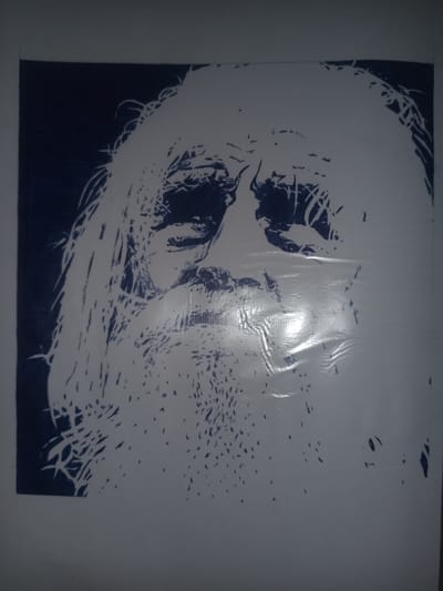 The image is a close-up, head-and-shoulders portrait of an elderly man drawn in a stencil style with dark blue ink on a white background. The man has a long, unkempt beard and long, flowing hair. His facial features are rendered with stark contrasts, emphasizing deep-set eyes, a prominent nose, and wrinkles. The drawing appears to be on a textured surface, possibly a large sheet of paper or fabric, which exhibits some creasing and a shiny, reflective patch in the lower right quadrant, suggesting it might be under some lighting or plastic wrap. The background is mostly white, with a dark blue area on the upper left, possibly representing hair or shadow. The style is abstract, with many sharp, angular lines and irregular shapes used to define the form. The image is not located in Gulbag, Uzbekistan, nor does it contain any contextual information about its location.