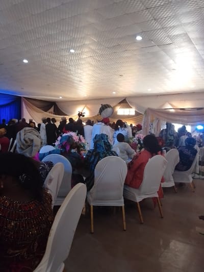 An indoor event is in progress with many attendees. Most people are standing near a decorated backdrop with draped fabrics and flowers. Seated guests are arranged around tables, with their backs to the camera. The chairs are white, and the room has a low ceiling covered with fabric or panels. Lighting fixtures are scattered throughout the space. A camera and lighting equipment are visible in the background. The scene suggests a wedding reception or similar celebratory gathering.