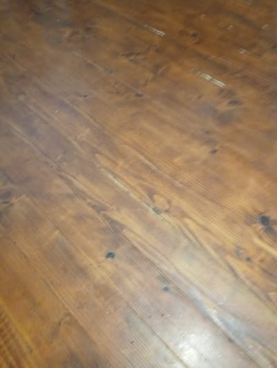 The image shows a close-up, angled view of a wooden floor. The floor is made of planks of wood that run diagonally from the bottom left to the top right of the frame. The wood appears to be stained a medium-dark brown, with visible grain patterns and knots. Some areas of the floor are lighter, suggesting wear or different wood types. There are a few small, dark spots on the surface, possibly imperfections in the wood or dirt. The lighting creates some reflections and highlights on the glossy finish of the wood. The image does not contain any people, objects, or actions, and it is not possible to determine its location within Verkhniachka, Ukraine.