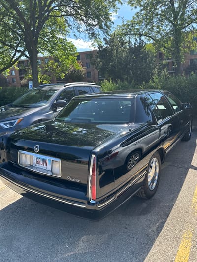 The image shows a black Cadillac DeVille parked on a paved street. The license plate reads "GRUVN" and indicates that the vehicle is registered in Illinois. The car is parked near a curb, with a yellow line visible on the pavement. In the background, there are trees and a multi-story brick building, suggesting an urban or suburban environment. The photo appears to have been taken in Arlington Heights, USA.