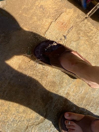 The image depicts a top-down view of two individuals' feet on a light-colored, textured stone or concrete surface under direct sunlight. A stream of water is actively flowing from the upper right, hitting a dark brown flip-flop or sandal worn on a light-toned foot. Water splashes are visible around this foot and the sandal, creating wet patches on the ground. A small reddish mark is present on the visible ankle/lower shin of the foot being rinsed. In the lower right corner, the toes of a second foot, wearing a brown sandal and featuring a silver toe ring on the second toe, are visible. A prominent, elongated dark shadow, likely from a person or an object, extends across the left side of the frame, indicating strong illumination. The scene is located in Πλατύς Γιαλός, Greece.