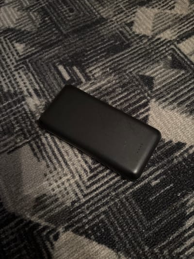 A black power bank is resting on a dark, patterned carpet. The carpet features geometric designs in shades of gray and black. The power bank is rectangular with rounded corners and appears to have three small indicator lights on its surface. The image is taken from a top-down perspective, with the power bank positioned slightly off-center. There are no people or any other objects visible in the scene. There is no information to determine the location in Chernogorsk, Russia.