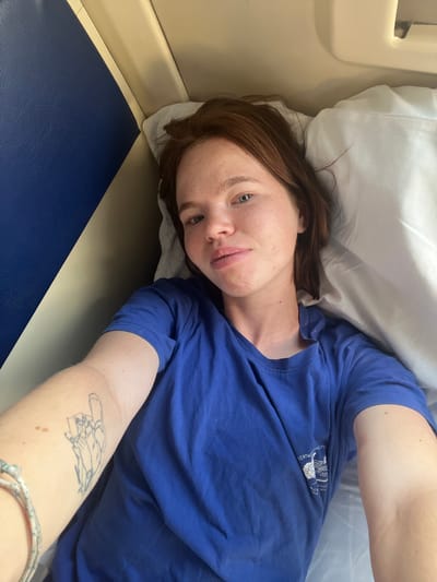 A young woman with reddish-brown hair lies on a pillow in what appears to be a train or bus compartment. She is wearing a blue t-shirt and looking at the camera. Her left arm is visible, showing a tattoo of a figure. The compartment has blue and beige upholstery.