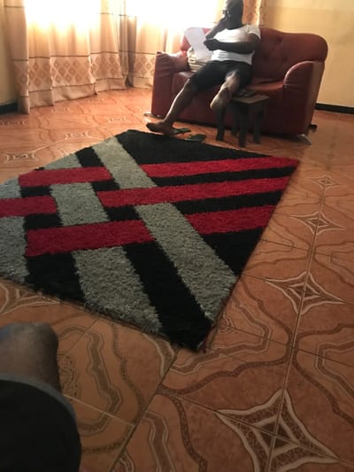 A man sits in a red chair, holding a white document. Next to him is a small wooden table. A multi-colored rug with black, red, and grey stripes lies on a tiled floor with a geometric pattern. Light enters from the side, illuminating a patterned curtain. A dark shape is visible in the bottom right corner.