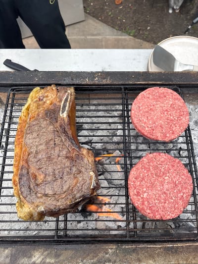 Two raw hamburger patties and a large cooked steak are placed on a charcoal grill. The charcoal is hot enough to produce flames. A spatula is next to the grill. A leg is visible in the lower left. The location is not determinable from the image.