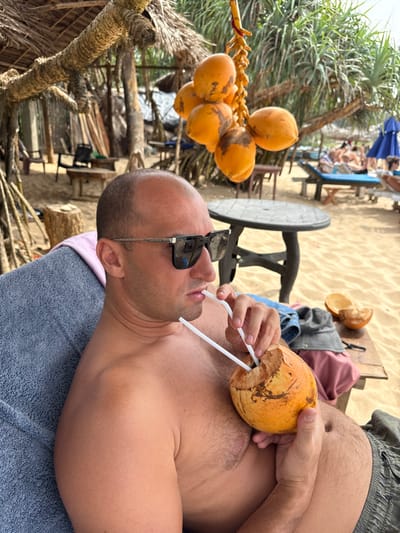 A bald man wearing sunglasses lies on a towel, shirtless, and drinks from a coconut with two straws. Several other coconuts hang from a nearby tree. In the background, other beachgoers are visible on lounge chairs. The scene appears to be a sunny beach setting with sand and tropical vegetation.