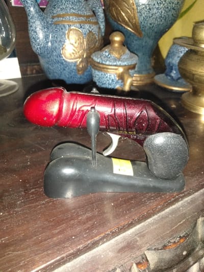 The image displays an interior setting in Косшы, Kazakhstan. A novelty lighter, designed to resemble a phallus, is centrally positioned on a dark brown wooden surface. The lighter features a glossy red, dark red, and black finish with textured patterns on its body, a black handle, and a silver trigger, resting on a black stand. The wooden surface exhibits minor scratches, scuffs, and dust. In the blurred background, several decorative objects are visible: a blue ceramic teapot with gold embellishments, a smaller blue ceramic lidded container, a tall blue ceramic vase with a large gold leaf design, a brass-colored item, and a clear glass object. No individuals are present in the scene.