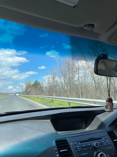 The image is taken from inside a vehicle, capturing a view through the windshield. Outside, a road stretches into the distance, bordered by a metal guardrail and a grassy area with yellow flowers. Beyond the road, leafless trees fill the horizon under a blue sky dotted with white clouds. Inside the vehicle, the rearview mirror is visible, along with the dashboard and radio console of the car. The radio console shows buttons and labels such as "FM," "AM," "CD," "AUX," "VOL," "CLOCK," and "ANTI-THEFT." An air freshener is hanging from the rearview mirror. The location, Stareishino, Bulgaria, is not confirmed by the image.