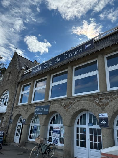 The image shows the exterior of a building located at 48.633 latitude and -2.051 longitude in the city of Dinard, France. The building is made of stone and has a sign above the entrance that reads "Yacht Club de Dinard". There are large windows on the upper floor and several arched doorways on the ground floor. The doors are white and have glass panels. There is a bicycle parked in front of the building. The building is set against a blue sky with white clouds. The sky also contains a rainbow-like formation near the top of the image. A sign above the doorway reads "RESTAURANT & BAR ROOFTOP YACHT". There is another sign advertising "DINARD YACHTING" on the right side of the building. A "EF Voile" sign is visible on the far right side of the building. The sign has a picture of a sailboat and says "Ecole de Voile."